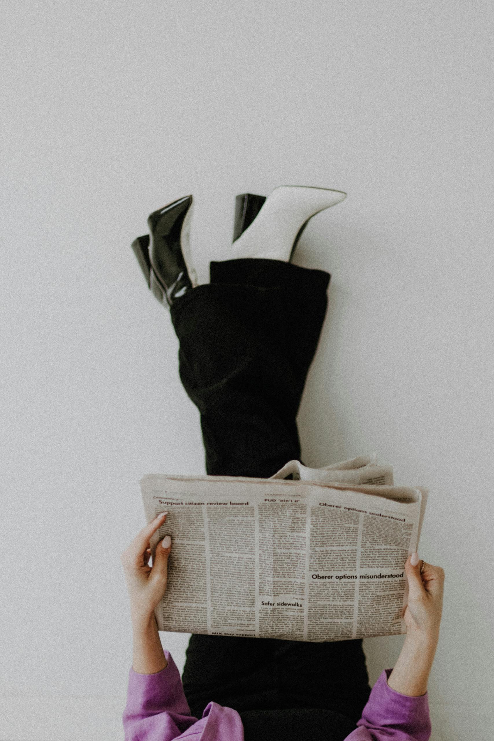 Fashionable woman reading newspaper with legs up against a wall in a studio setting.